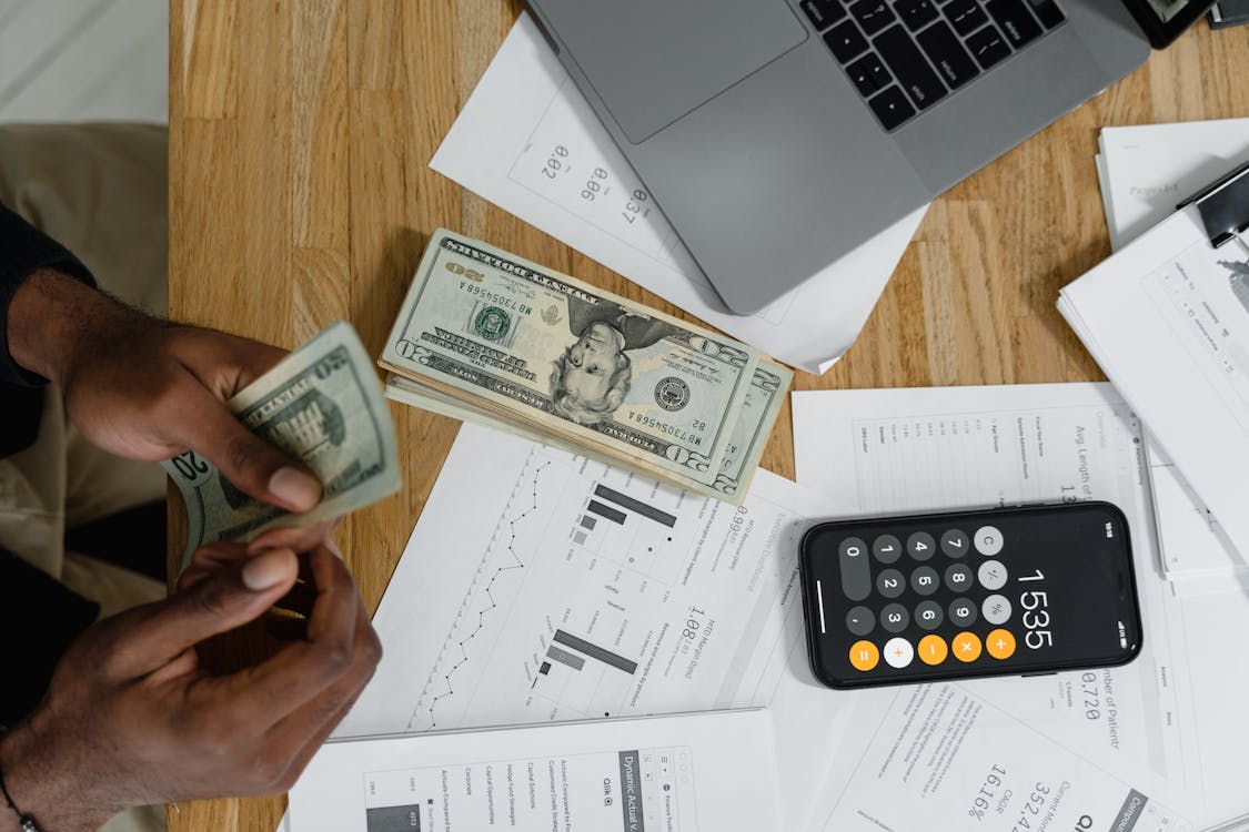 Person counting money at a desk with calculator, laptop, and financial reports